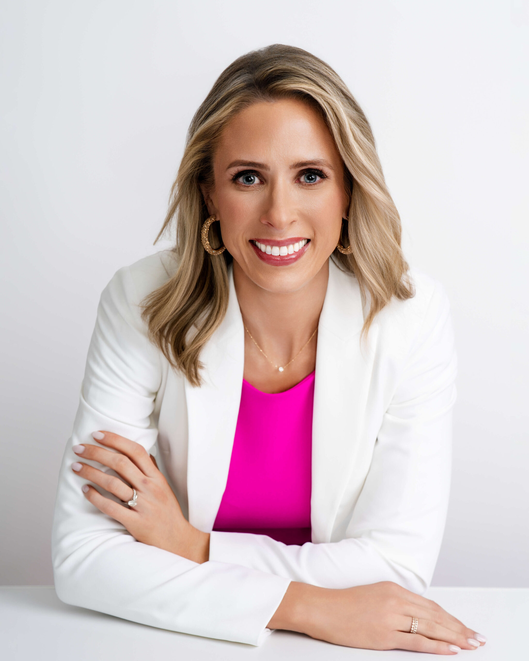 Modern close-up branding headshot of a blonde woman in a pink dress and white blazer, smiling with hands crossed on a white table.