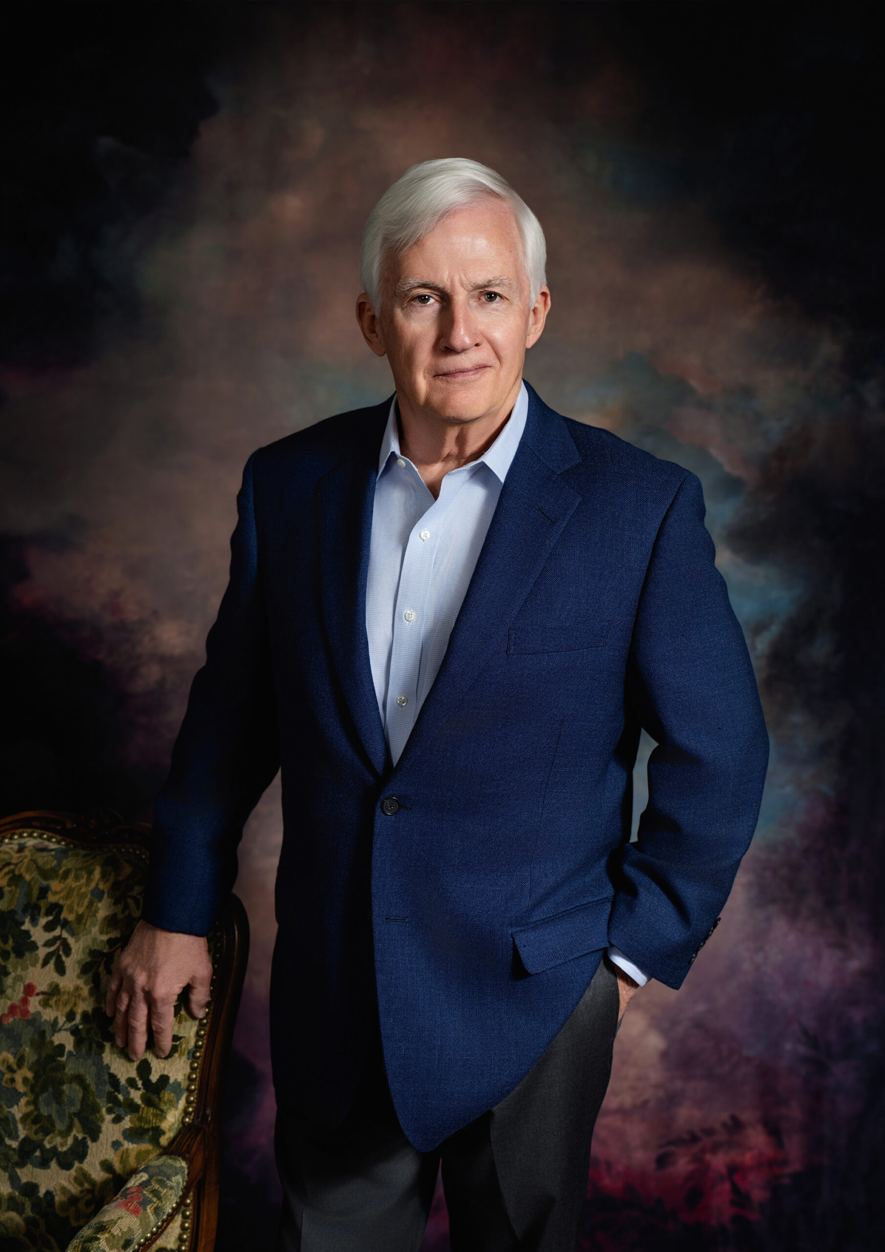 Distinguished older man in a tailored navy blazer and light blue dress shirt posing for a professional headshot on a painterly fine art backdrop
