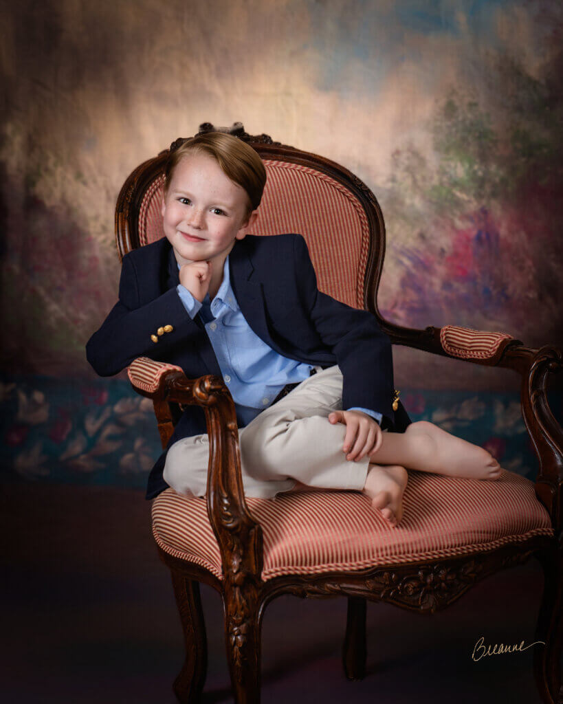 Six-year-old boy with freckles in a navy blazer sitting cross-legged in an antique French armchair during an heirloom children's portrait session near Mountain Brook, Alabama