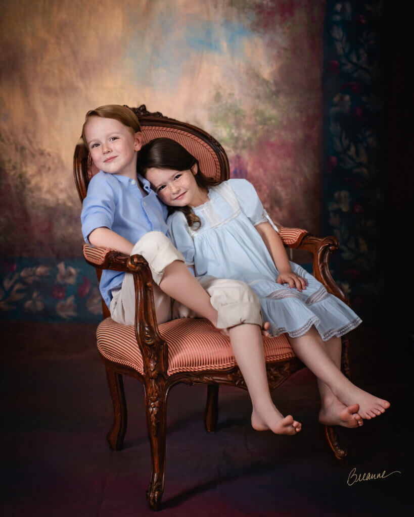Brother and sister with freckles leaning together in an antique striped armchair during a sibling heirloom portrait session in Birmingham, Alabama