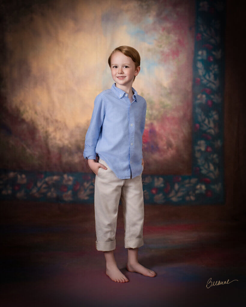 Seven-year-old boy with freckles in a blue oxford shirt and rolled khakis standing barefoot in front of a hand-painted backdrop in a Trussville, Alabama portrait studio