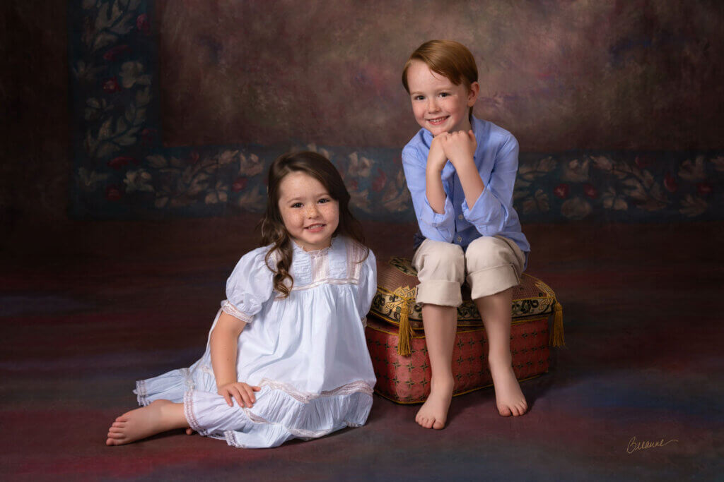 Brother and sister with freckles sitting together on a French cushion in front of a painterly landscape backdrop for an heirloom sibling portrait